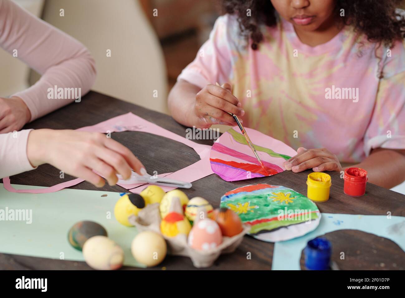 Petite fille de race mixte sérieuse avec paintbrush paint papier oeuf de Pâques tout en étant assis près d'une table en bois à côté de la jeune femme aidant elle Banque D'Images