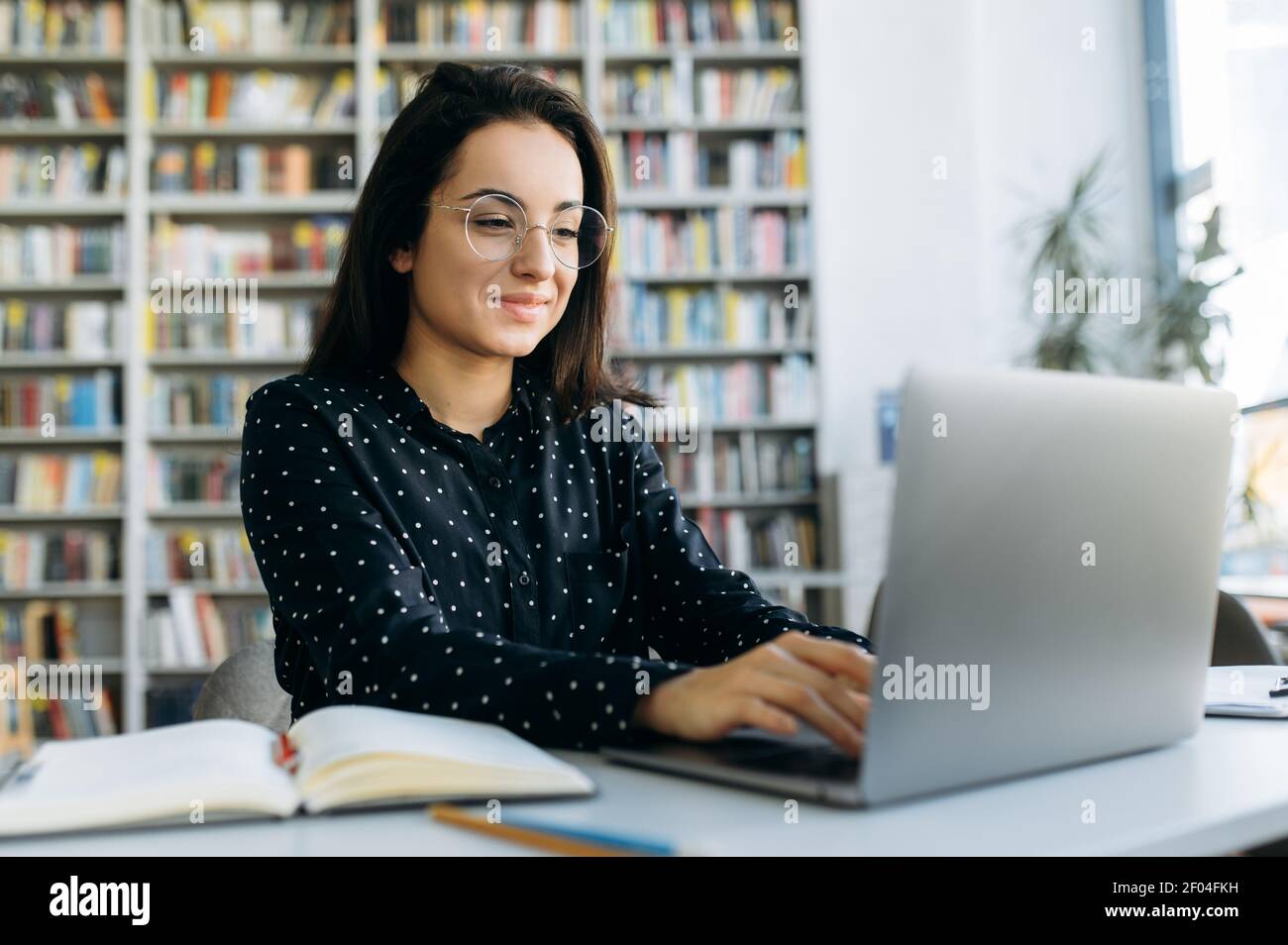 Une femme très attirante sur le lieu de travail. Jolie femme utilisant un ordinateur portable, développant un nouveau projet ou une nouvelle application, assis à la table dans un bureau moderne, sourit Banque D'Images