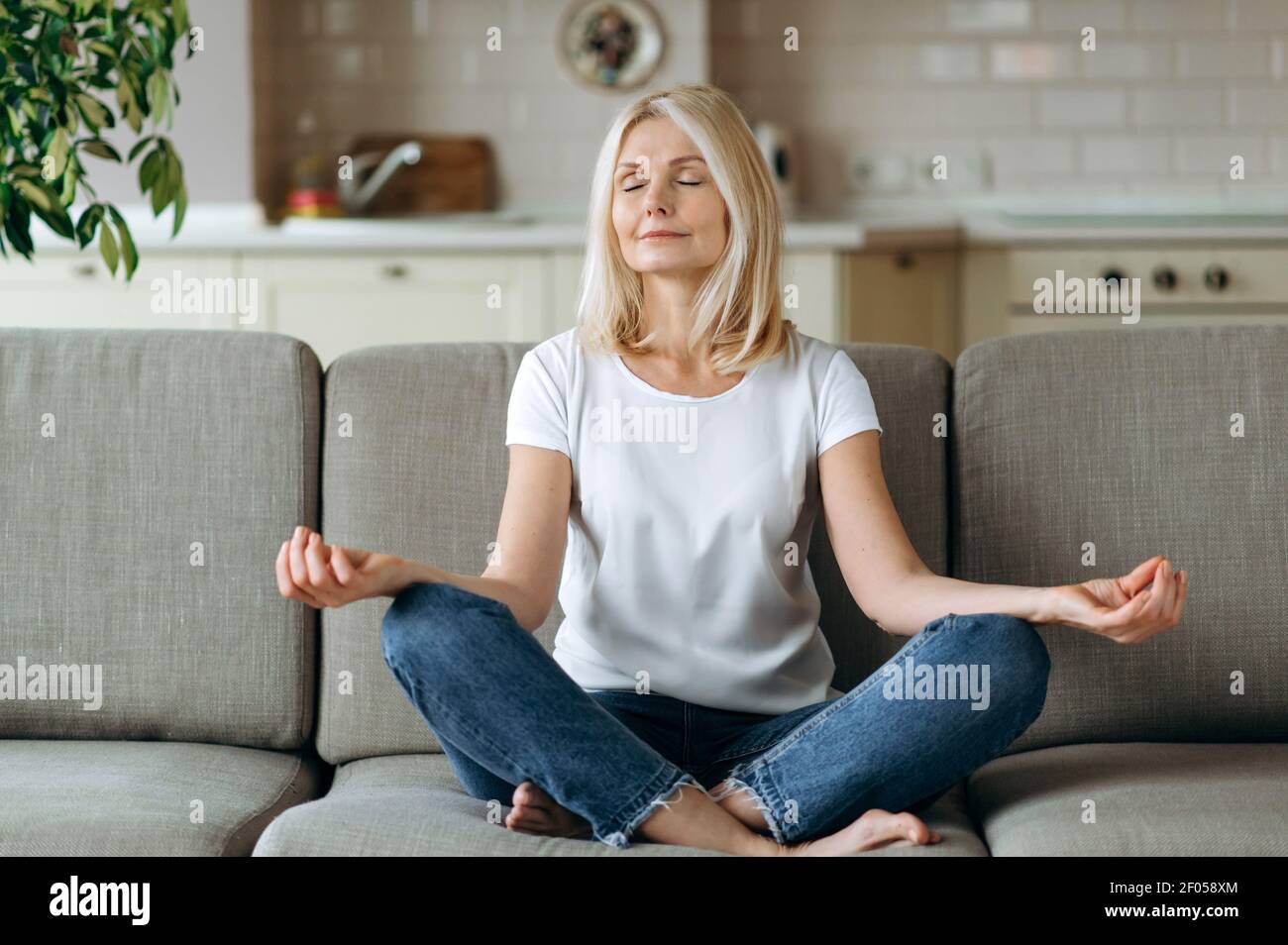 Calme et attrayante femme d'âge moyen assis sur un canapé à la maison et de refroidissement. La femelle senior pratique le yoga et la méditation en position de lotus avec les yeux fermés, soulagent le stress, concept de calme Banque D'Images