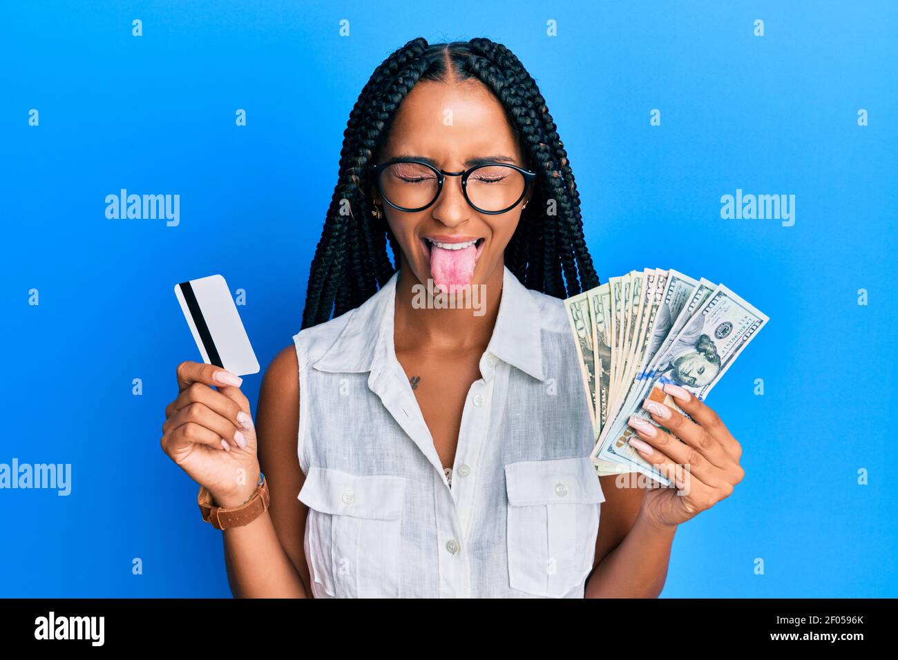 Belle femme hispanique tenant des dollars et carte de crédit collant la langue dehors heureux avec l'expression drôle. Banque D'Images