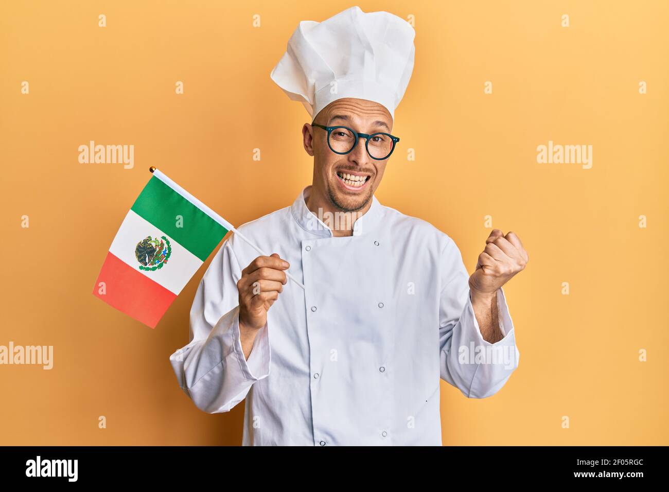 Homme chauve avec barbe portant un tablier de cuisinier professionnel tenant le drapeau du mexique criant fier, célébrant la victoire et le succès très excité avec le bras levé Banque D'Images