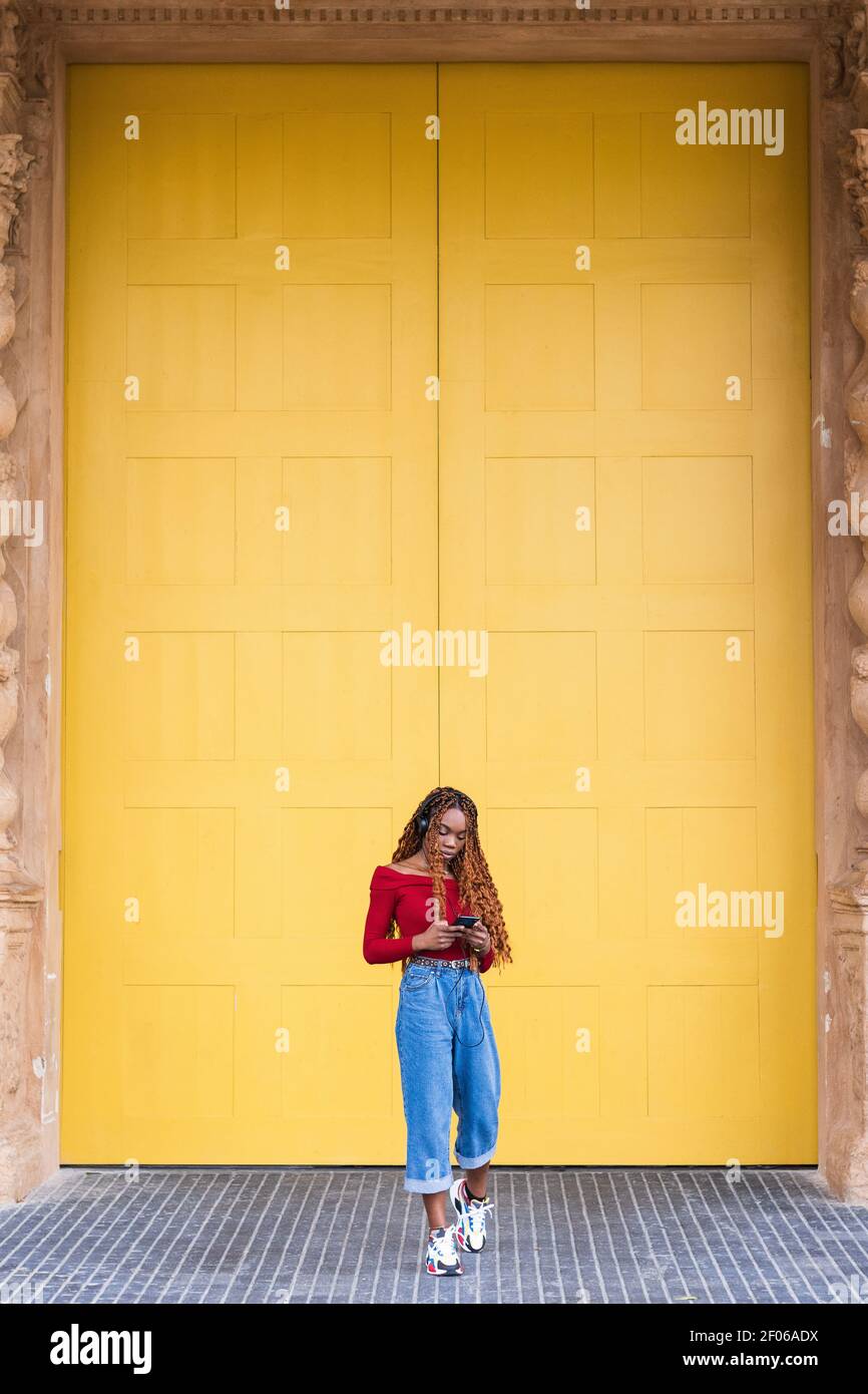 Femme afro-américaine debout près d'énormes portes en bois pendant qu'elle est sur entrée du bâtiment avec téléphone portable pour écouter de la musique casque Banque D'Images