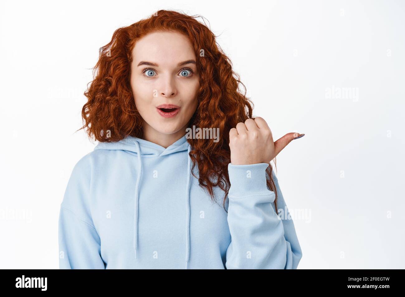 Génial. Impressionnée fille de gingembre avec des cheveux bouclés, look excité, pointant le doigt vers la droite et souriant étonné, port de sweat à capuche, fond blanc Banque D'Images