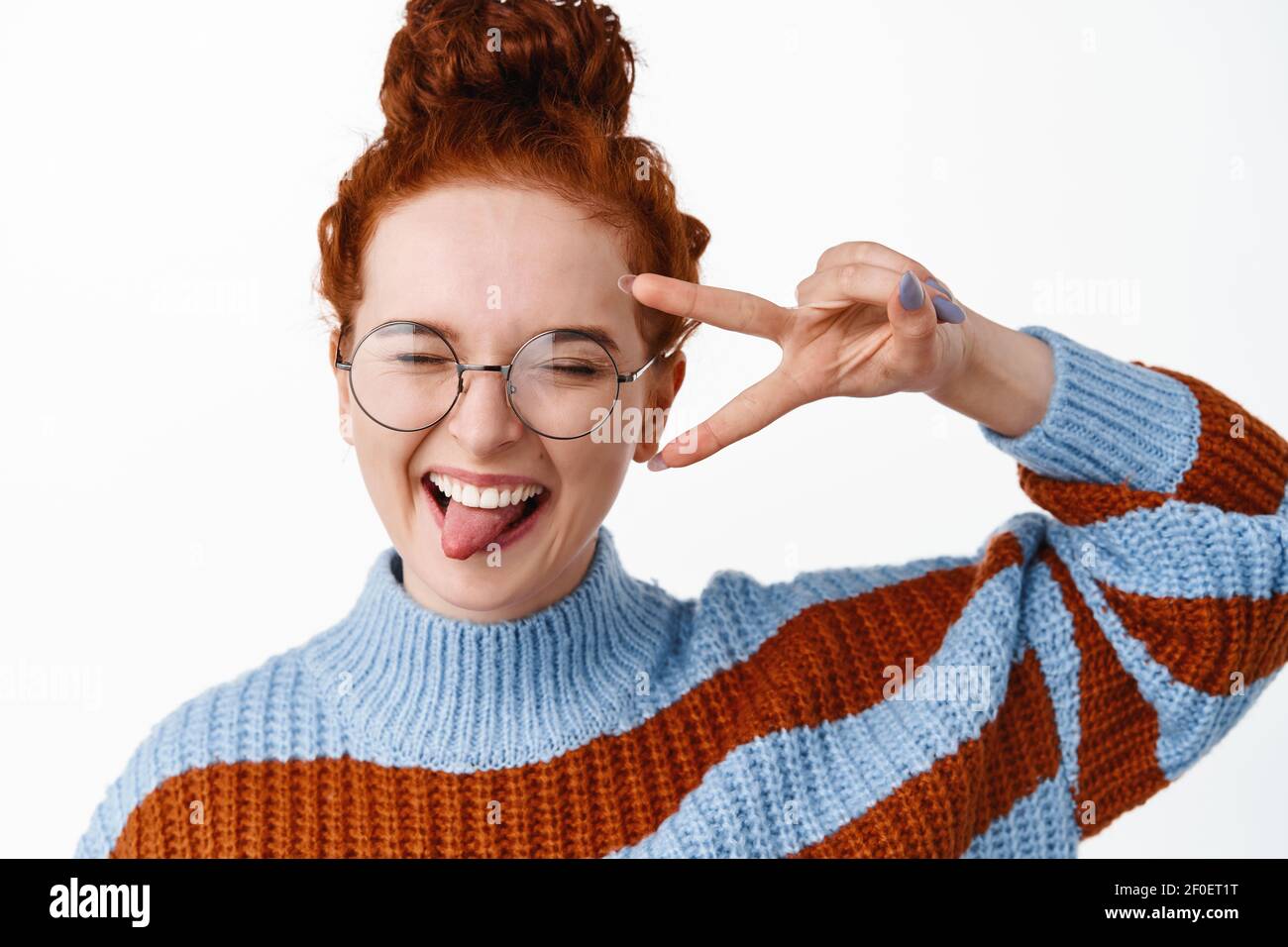 Gros plan portrait d'une fille heureuse et positive à tête rouge en lunettes, montrant la langue avec le V-signe et ayant plaisir, exprimer la joie et l'excitation, blanc Banque D'Images
