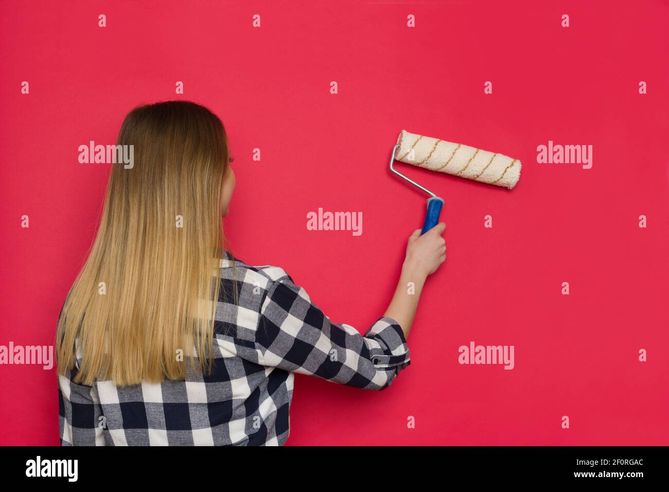 Jeune femme décontractée dans le maillot Lumberjack est peinture mur rouge avec le rouleau de peintre. Vue arrière. Prise de vue à la taille. Banque D'Images