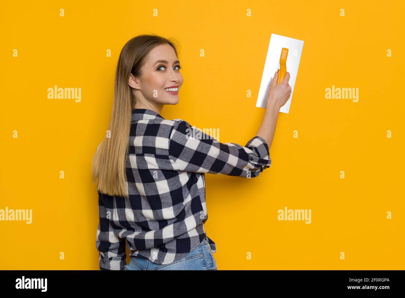 Jeune femme Blonde dans le maillot Lumberjack pose avec la truelle de finition sur le mur jaune et regardant au-dessus de l'épaule. Vue arrière. Prise de vue à la taille. Phot de concept Banque D'Images