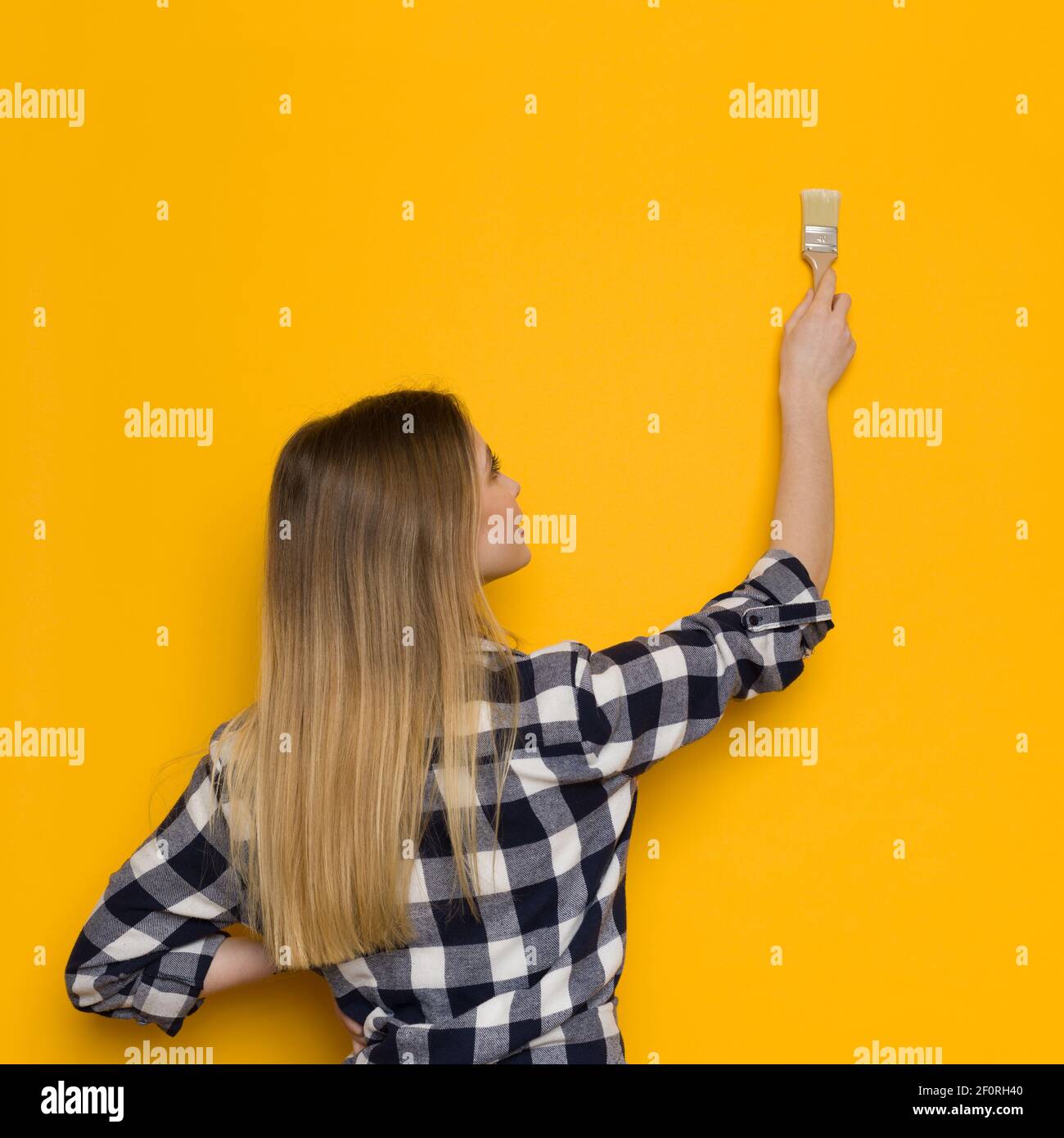 La jeune femme blonde en chemise de bûcherons peint un mur jaune. Vue arrière. Taille haute. Banque D'Images