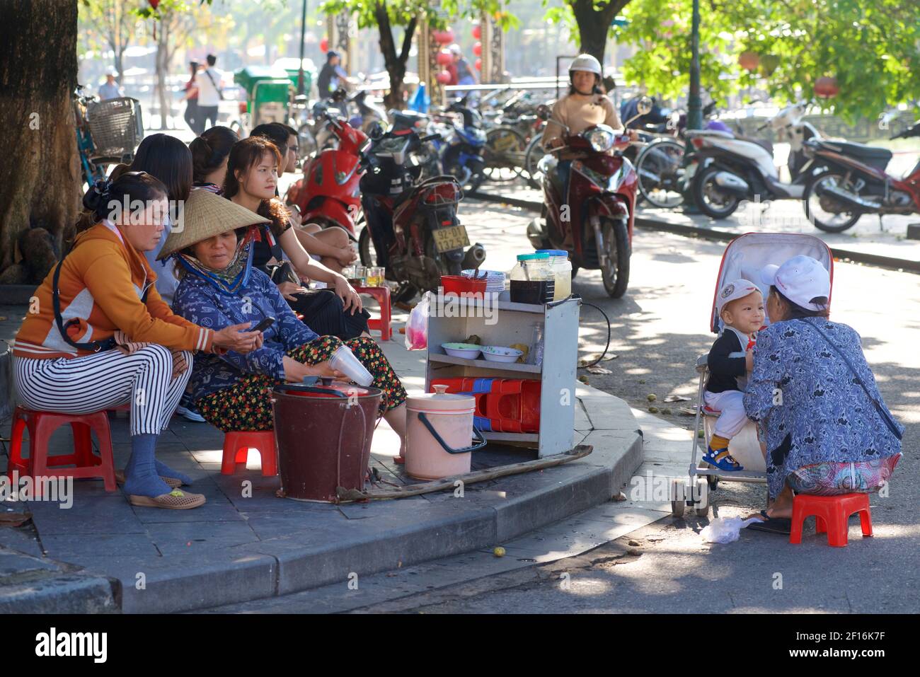 Vie de rue vietnamienne. Vendeurs vendant des collations sur le front de mer, Hoi an, Vietnam. Mère allaitant garçon en pram. Banque D'Images