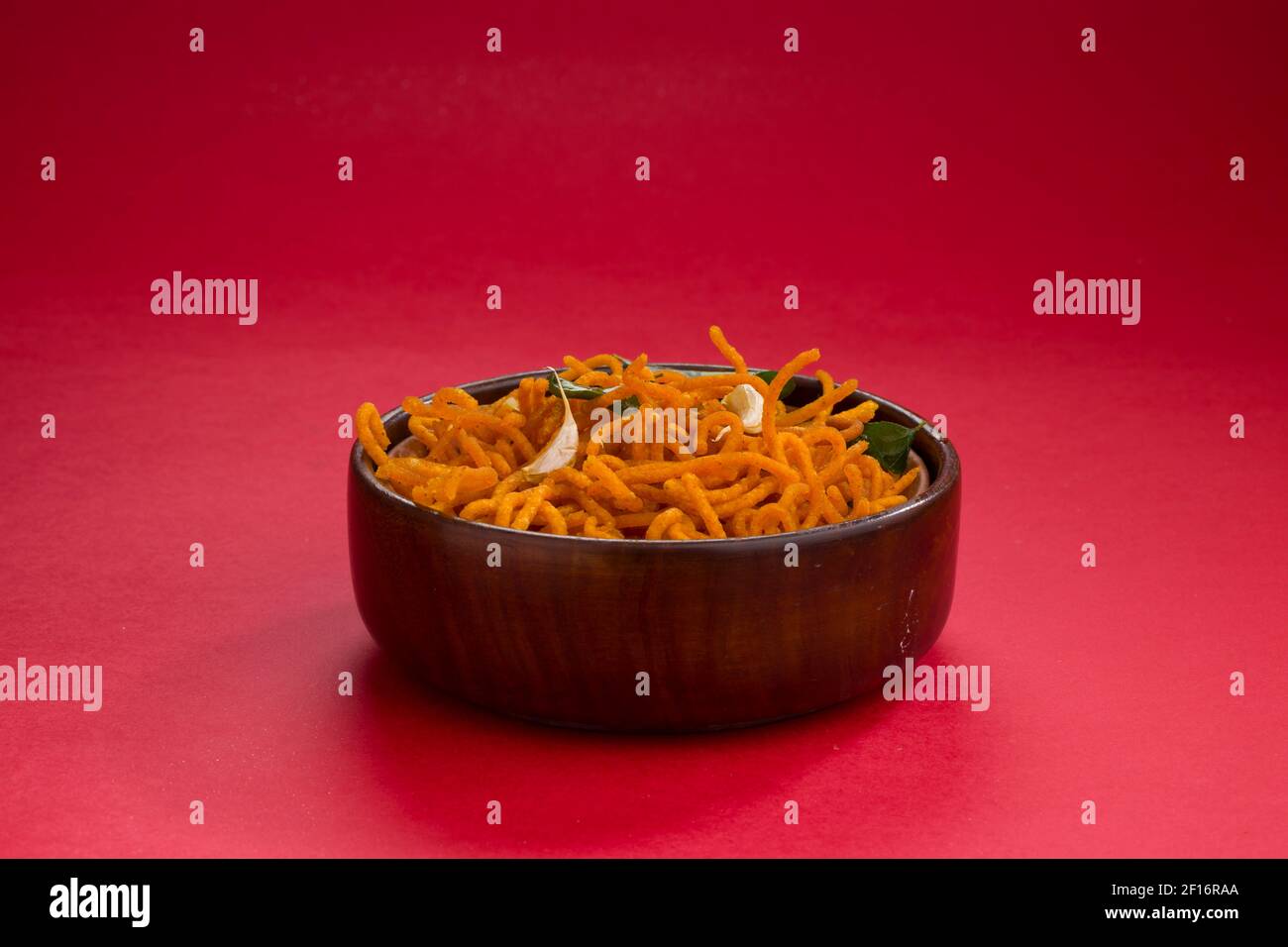En-cas indiens Besan sev ou Namkeen sev garni de feuilles de curry frites et de gousses d'ail dans un bol en bois à fond texturé rouge. Banque D'Images