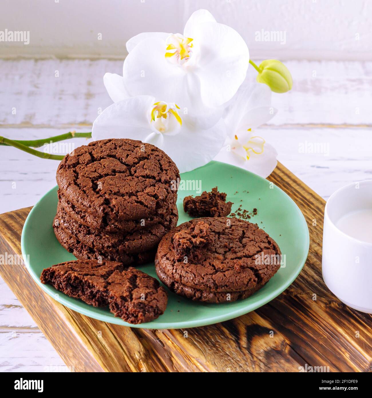 Petits gâteaux au chocolat pour le petit déjeuner avec un verre de lait sur une table en bois blanc avec une fleur d'orchidée en fleurs. Banque D'Images