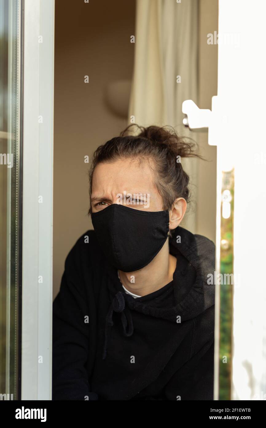 Un jeune homme avec un masque facial regardant par la fenêtre pendant son auto-isolement à la maison. Covid-19 corona virus isolement solitude concept Banque D'Images