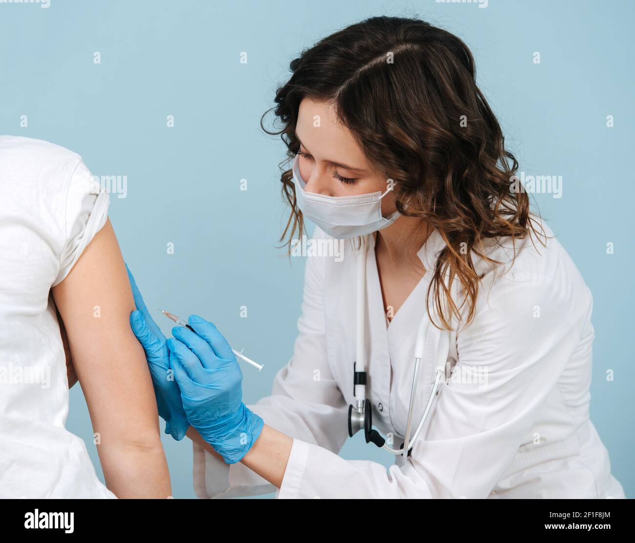 Infirmière soignante qui donne l'injection du vaccin de l'épaule. Sur fond bleu. Elle porte une robe blanche et un masque médical. Banque D'Images