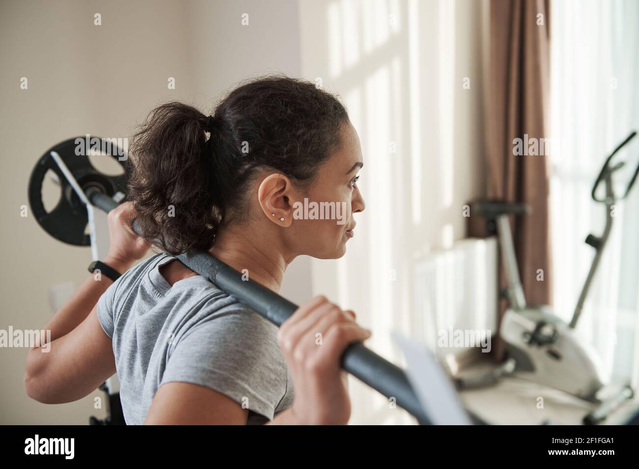 Vue arrière de la jeune femme sportive afro-américaine qui fait de l'exercice avec un squat lourd et une barbell dans la salle de gym à la maison. Entraînement lourd. Banque D'Images