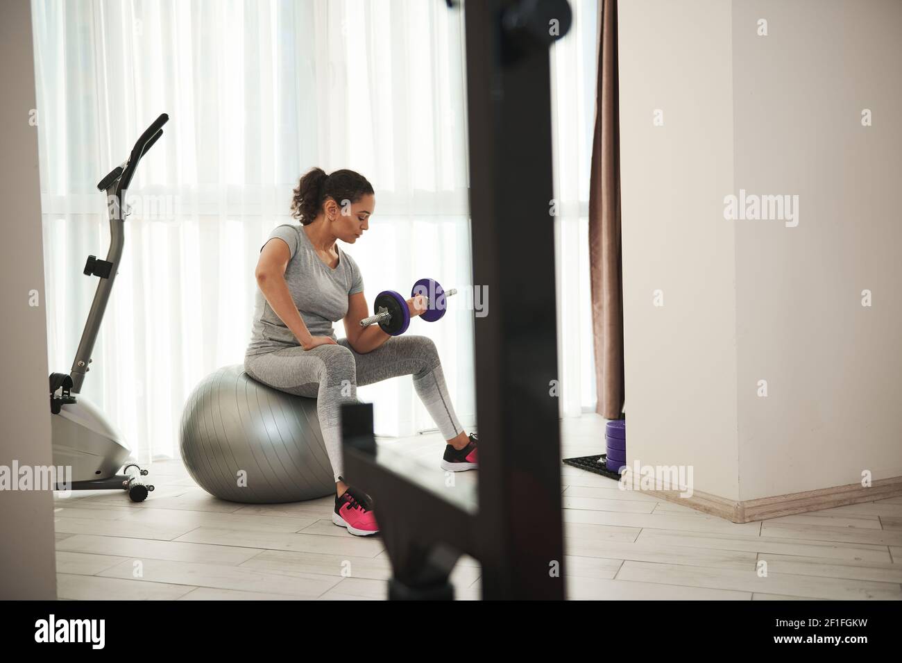Belle femme sportive pompant des biceps avec haltères tout en étant assise sur le ballon de fitness à la salle de gym à la maison. Entraînement à domicile. Banque D'Images