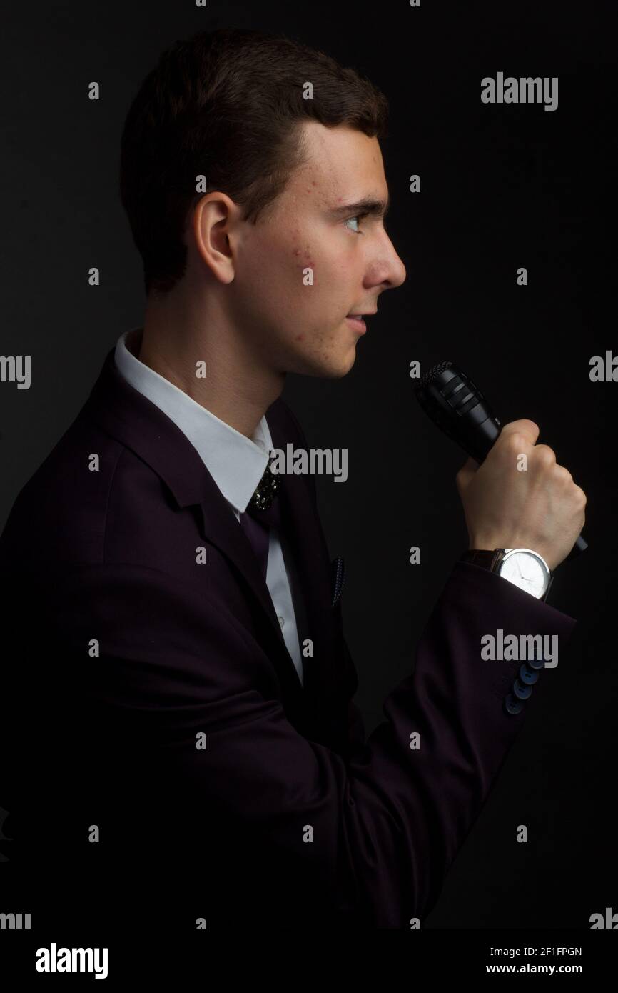 jeune homme aux cheveux sombres, vêtu d'une chemise blanche, d'un costume violet foncé et d'un noeud papillon en ruban, il tient un microphone dans les mains. Hôte ou chanteur-perform Banque D'Images