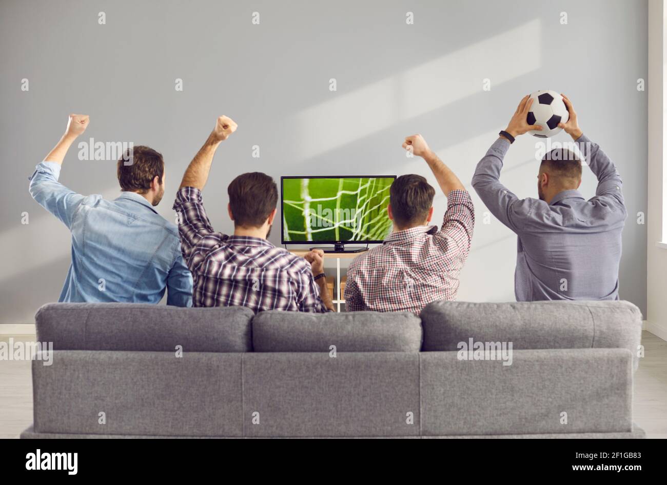 Quatre amis hommes adultes aux bras levés émotionnellement en regardant un match de football à la télévision à la maison. Banque D'Images