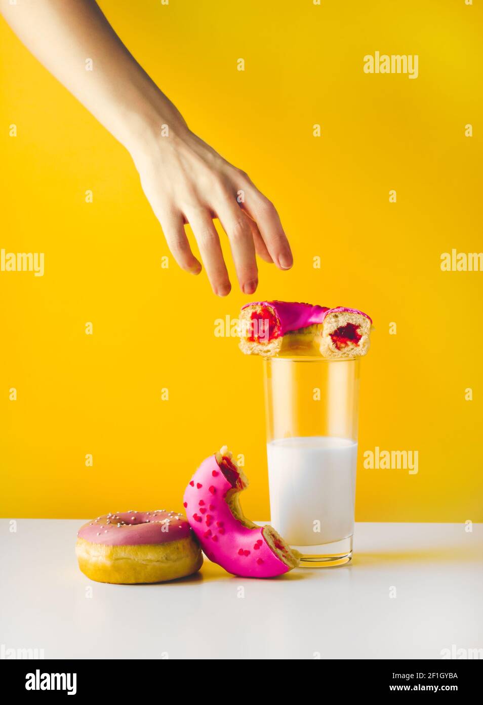 Femme caucasienne main atteignant des beignets délicieux avec confiture de baies de remplissage et de saupoudrage. Une pâtisserie rose et un verre de lait végétal sur fond jaune et gris Banque D'Images