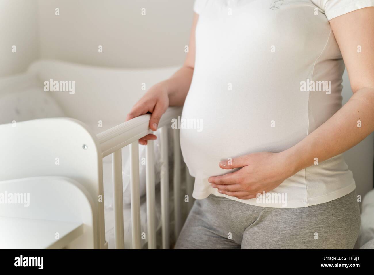 Gros plan sur la section médiane d'une femme enceinte du caucase debout inconnue par le socle dans une pièce lumineuse à la maison la tenant ventre - maternité et grossesse c Banque D'Images