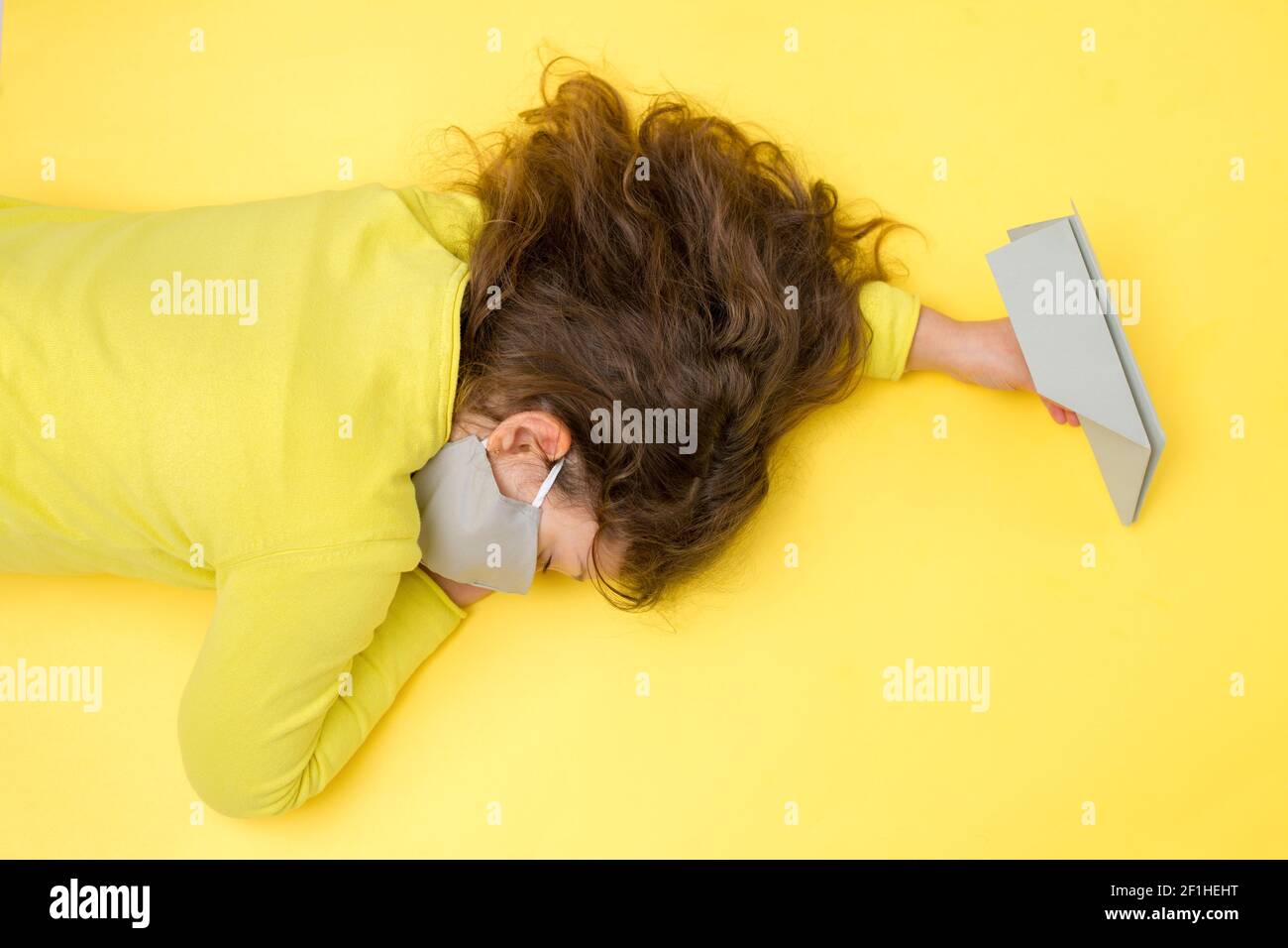 Une petite fille dormant dans un masque gris protecteur en main avec un avion en papier gris sur un fond jaune. Concept créatif de voyage, vacances pendant Banque D'Images