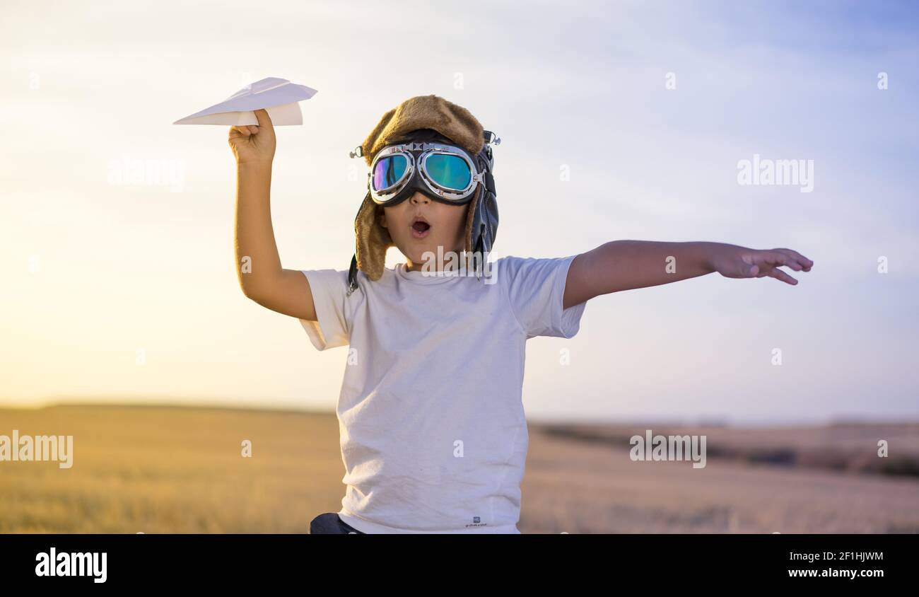 Little Boy wearing helmet et rêve de devenir un aviateur pendant la lecture d'un avion en papier au coucher du soleil Banque D'Images