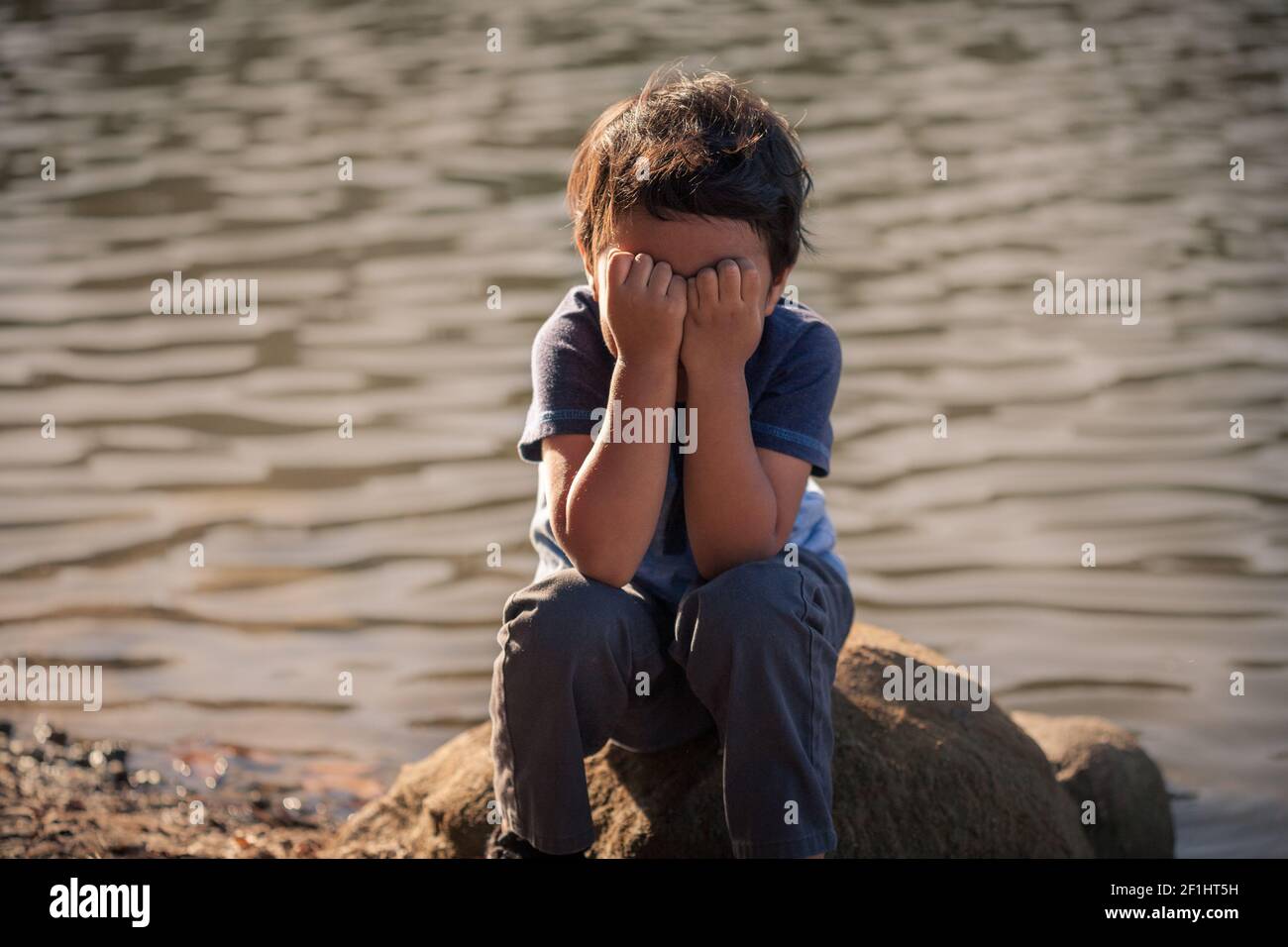 Un jeune garçon au bord d'un lac couvrant son visage, langage corporel exprimant des signes d'avertissement de violence émotive et d'isolement d'enfant. Banque D'Images