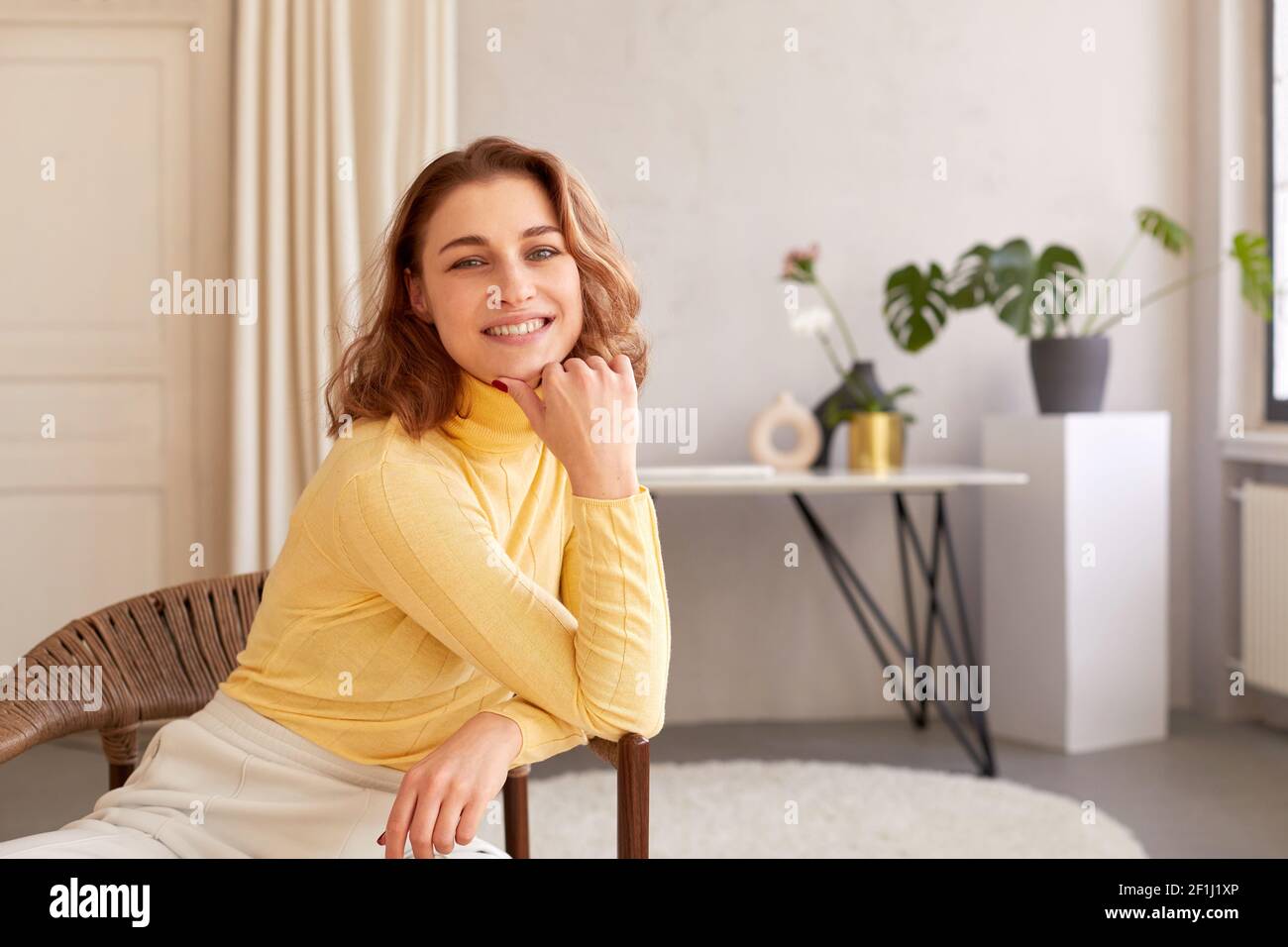 Content femelle avec un sourire charmant pendu sur la main et regardant à la caméra tout en étant assis sur une chaise en osier à la maison Banque D'Images