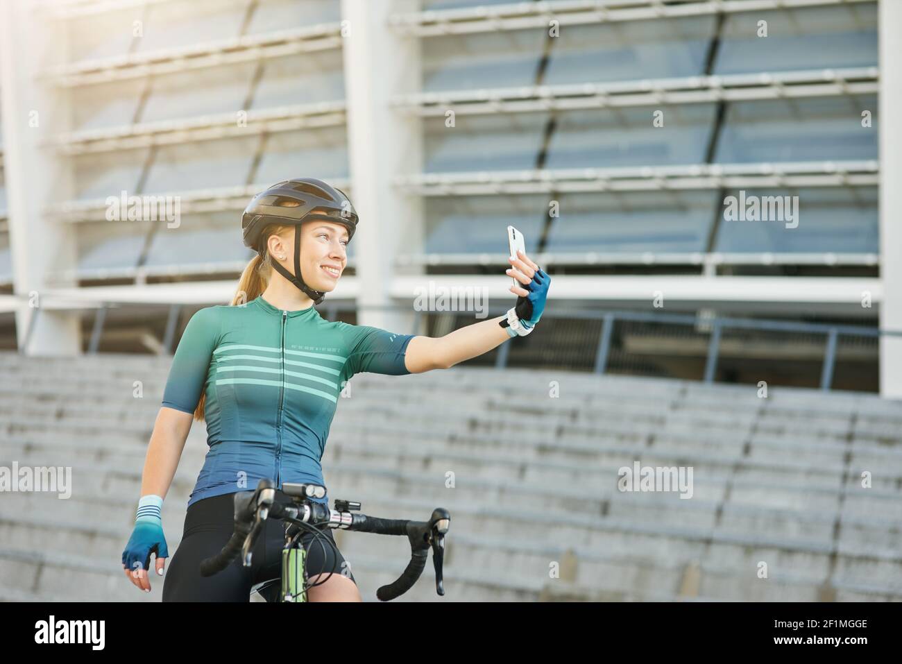 Adorable jeune femme, cycliste professionnelle souriant tout en tenant un smartphone et en prenant un selfie, debout avec son vélo à l'extérieur pendant la journée. Mode de vie actif, concept sportif Banque D'Images
