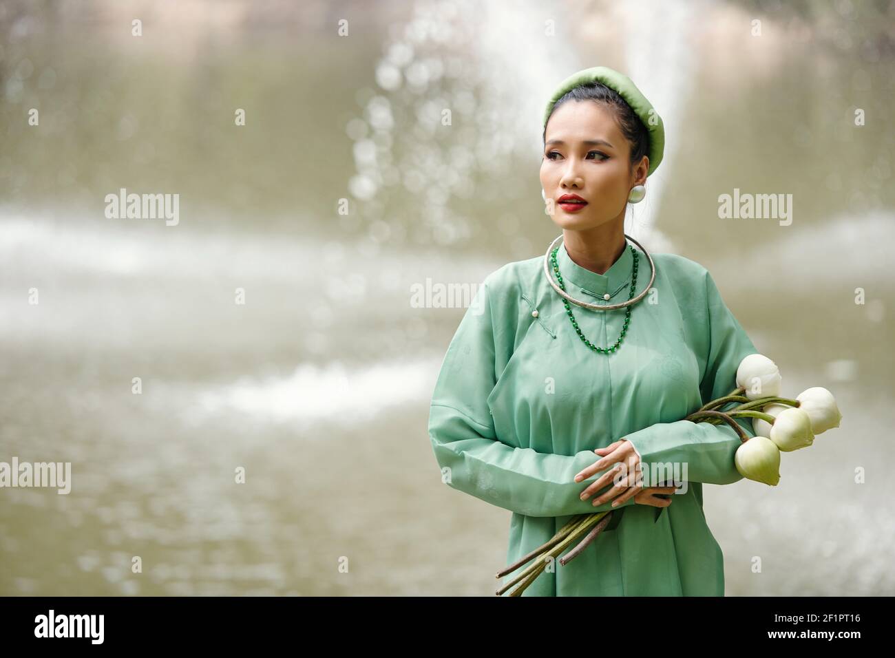 Portrait d'une jeune femme asiatique attirante dans la traditionnelle ao dai robe et serre-tête tenant des fleurs de lotus blanc et regardant loin Banque D'Images