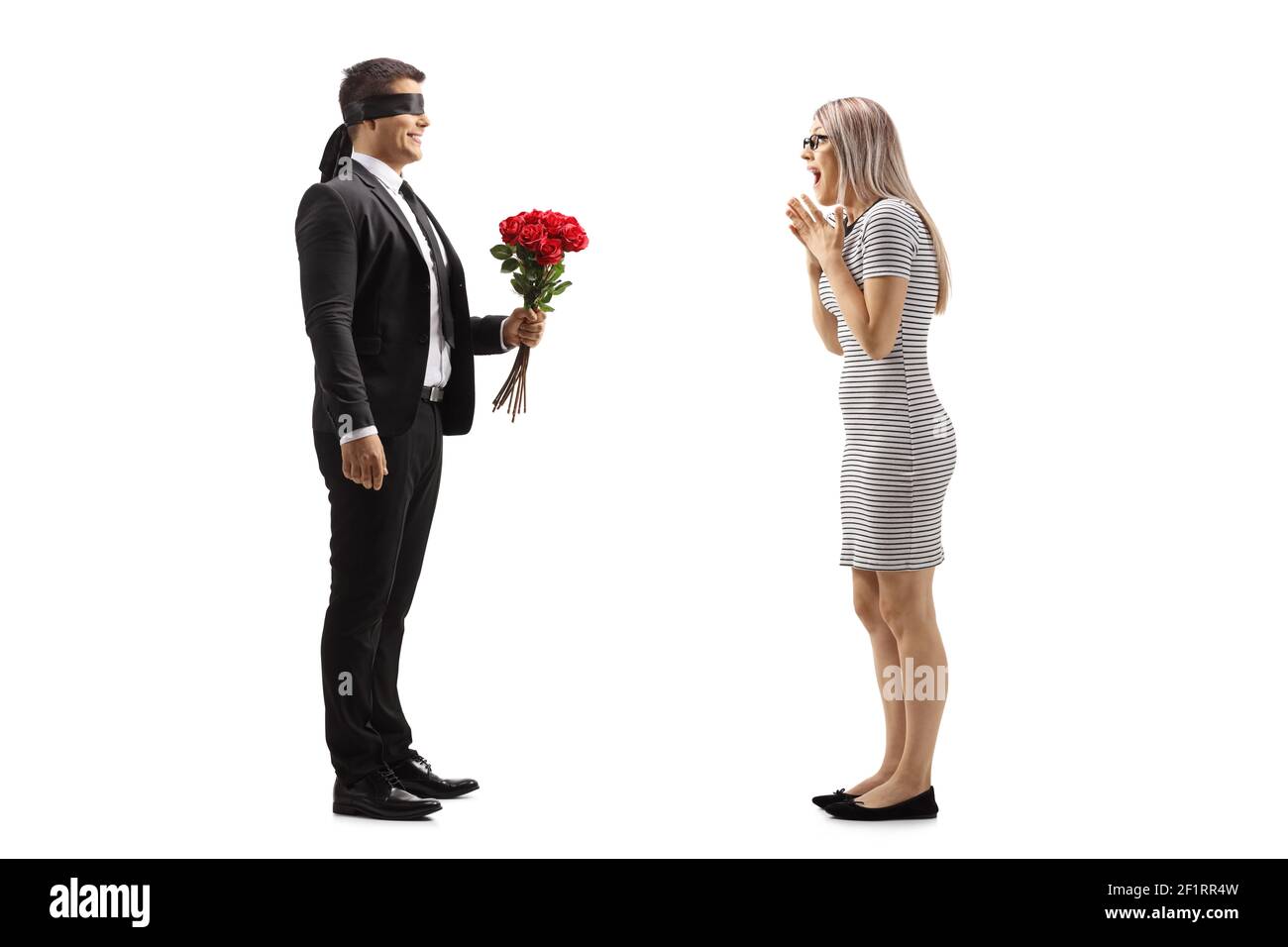 Photo pleine longueur d'un homme en costume avec les yeux bandés donnant un bouquet de roses rouges à un jeune femme excitée isolée sur fond blanc Banque D'Images