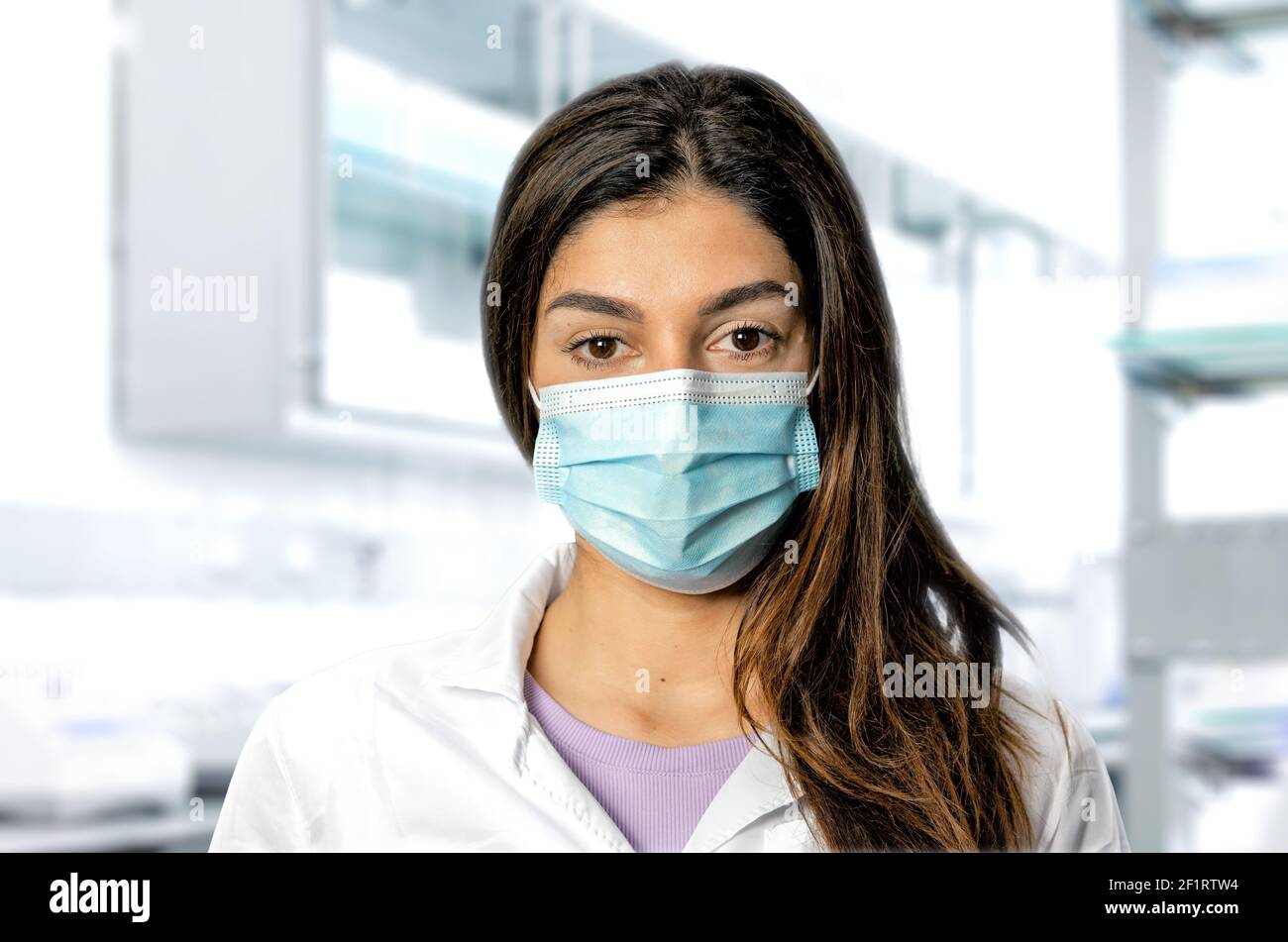 Un médecin brunette avec un masque chirurgical avec un laboratoire médical flou en arrière-plan. Une injection parfaite pour les pandémies, la recherche, les vaccins et la sécurité. Banque D'Images