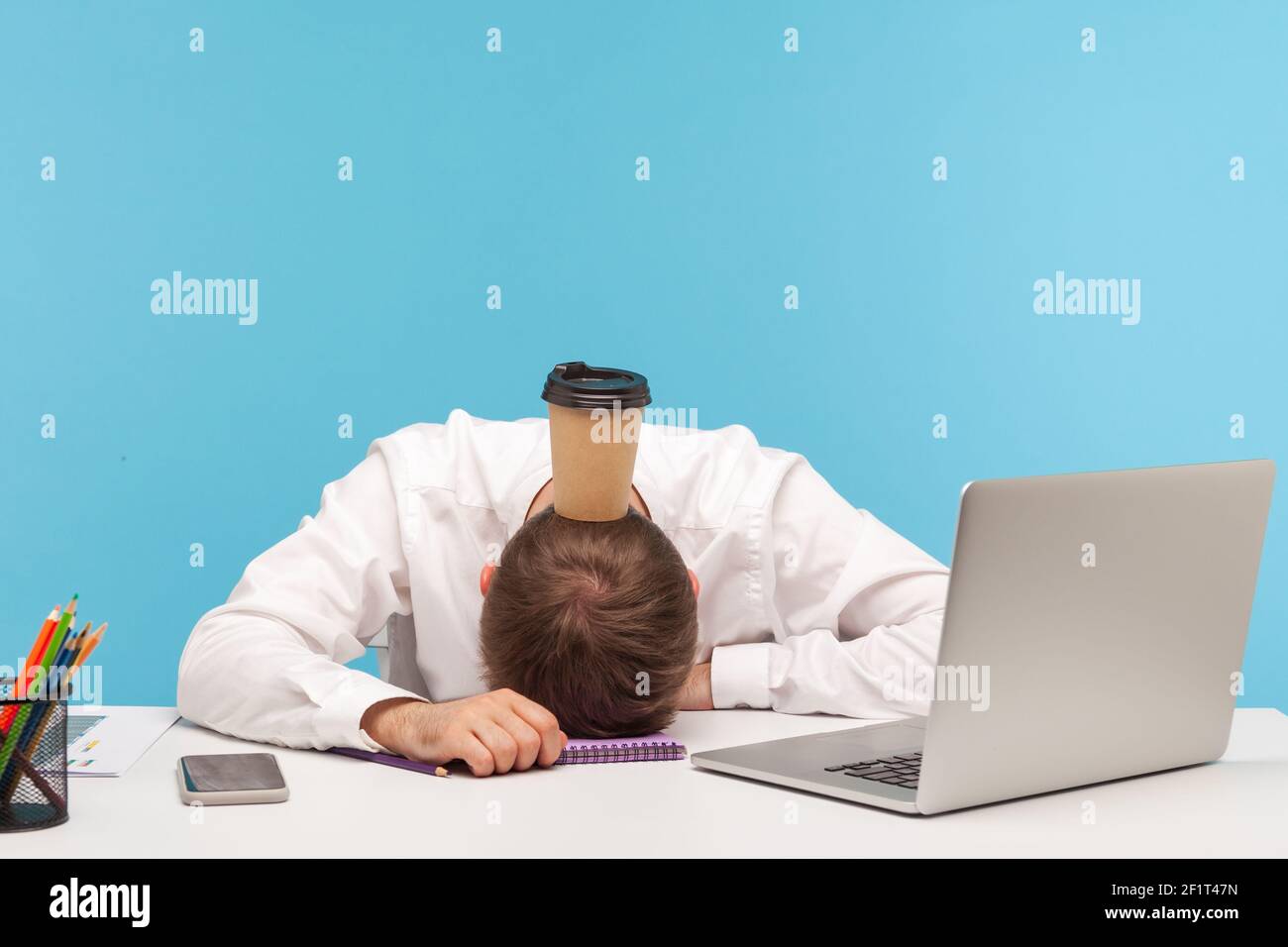 Travailleur de bureau épuisé surtravaillé allongé sur une table, tasse de café en papier debout sur sa tête, manque d'énergie, procrastination et professionnel burnout Banque D'Images