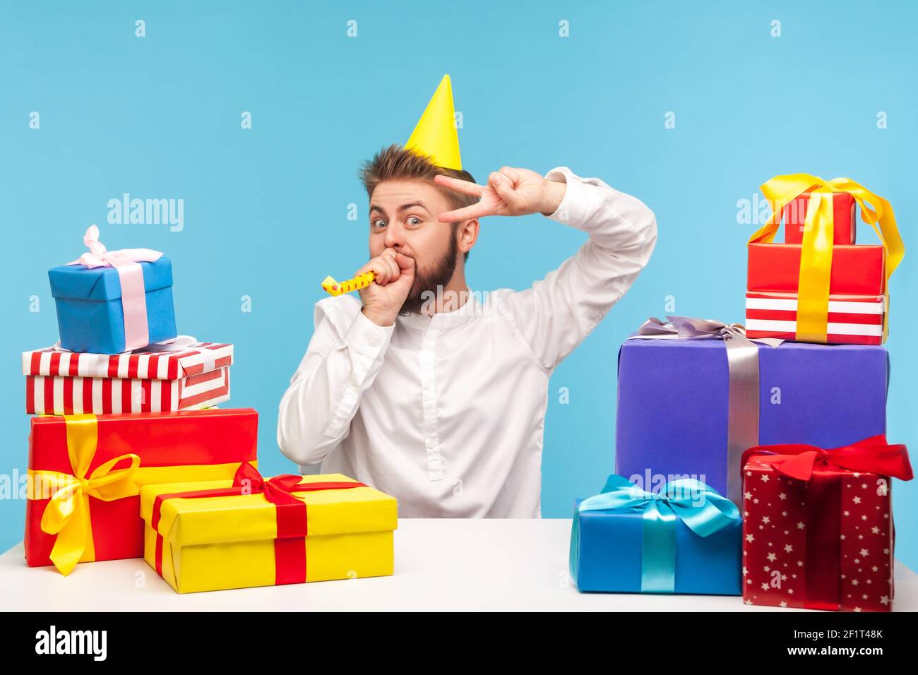 Un homme heureux dans un chapeau de cône soufflant dans le ventilateur de partie et montrant le geste de victoire assis sur le lieu de travail avec l'homme coloré boîtes cadeaux, agence de vacances. St. Intérieure Banque D'Images