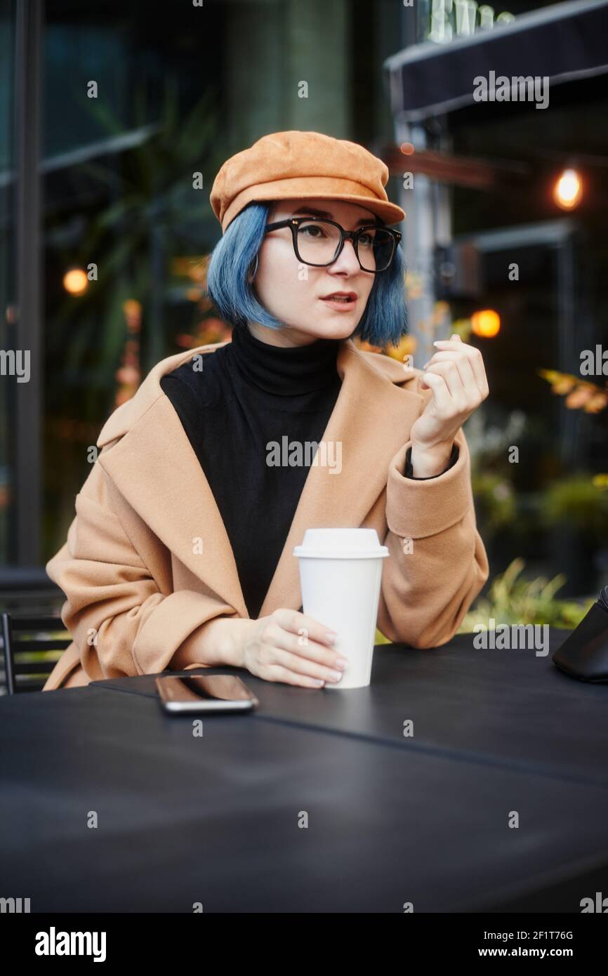 Une fille avec des cheveux bleus et des lunettes est assise à Une table dans un café dans la rue tenant un Tasse de café automne Banque D'Images