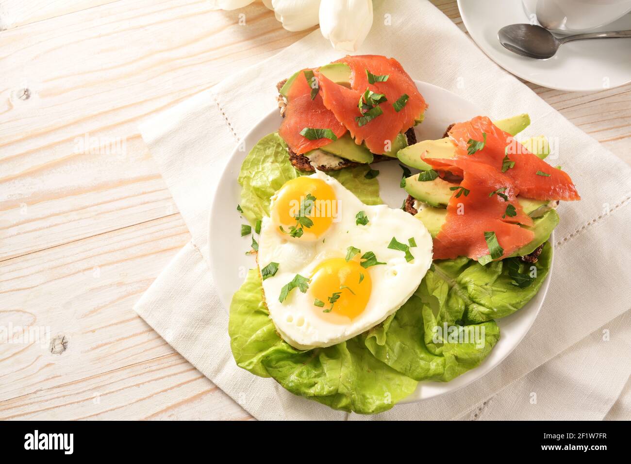 Deux œufs frits en forme de coeur, sandwichs avec avocat et saumon fumé sur salade de laitue, recette avec amour pour pâques ou Saint Valentin, bois blanc Banque D'Images