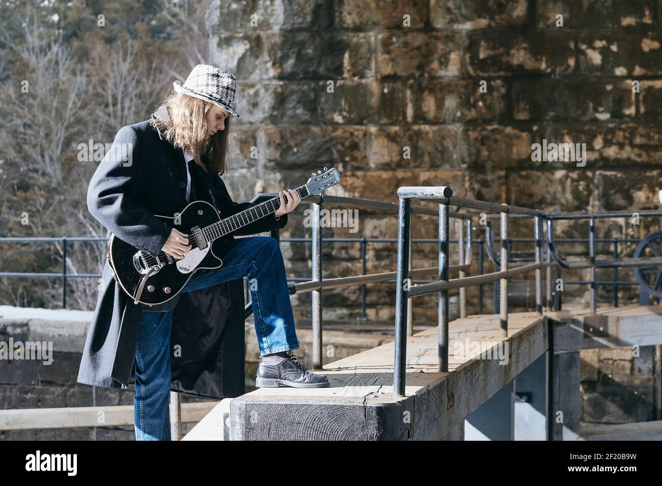 Jeune musicien masculin en manteau et chapeau debout sur les escaliers et jouer de la guitare électrique à l'extérieur du vieux bâtiment Banque D'Images