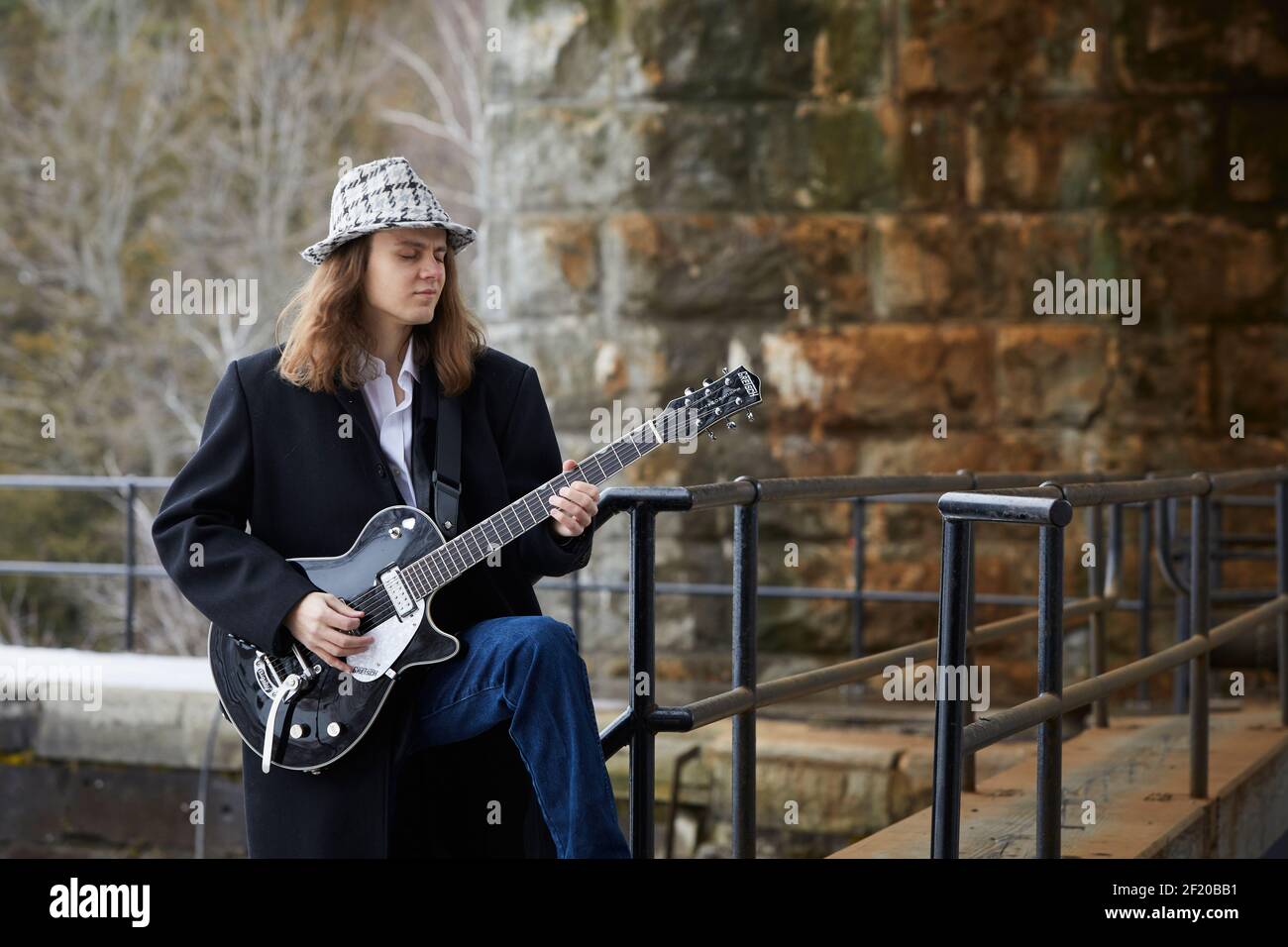 Jeune musicien masculin en manteau et chapeau debout sur les escaliers et jouer de la guitare électrique à l'extérieur du vieux bâtiment Banque D'Images