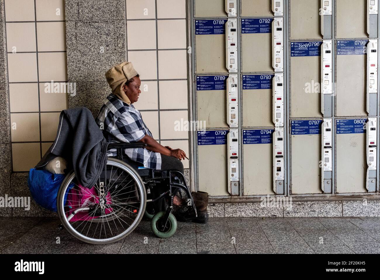Berlin, Allemagne. Femme de couleur sans domicile, tuant du temps et passant sa journée, assise dans son fauteuil roulant, garée devant les casiers à bagages du zoo Bahnhof. Banque D'Images