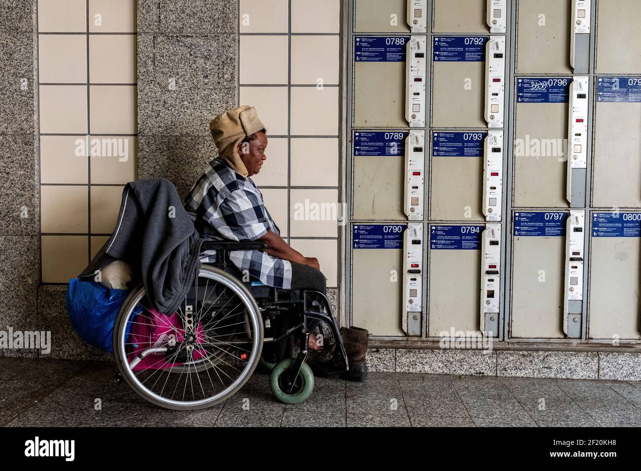 Berlin, Allemagne. Femme de couleur sans domicile, tuant du temps et passant sa journée, assise dans son fauteuil roulant, garée devant les casiers à bagages du zoo Bahnhof. Banque D'Images