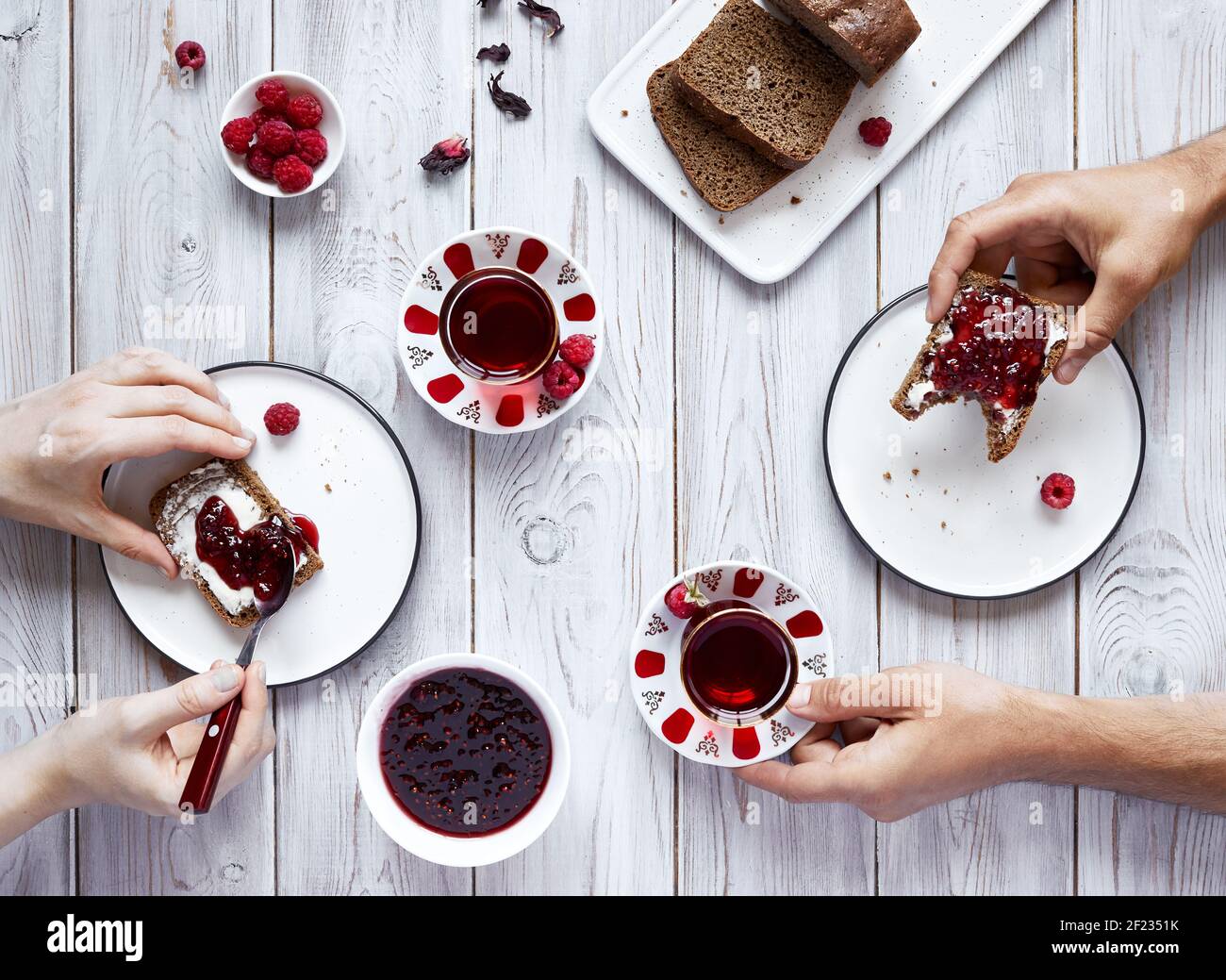 Homme et femme buvant du thé et mangeant des toasts à la framboise gelée au petit déjeuner sur une table en bois blanc Banque D'Images