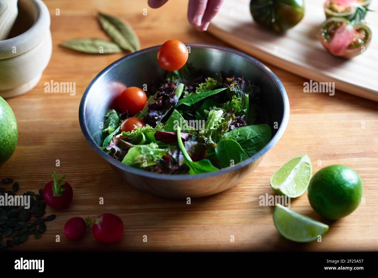 Une main jette une tomate cerise sur une salade dans un bol en métal sur une table en bois dans une cuisine. Dans la photographie gastronomique vous pouvez voir des radis, des graines, Banque D'Images