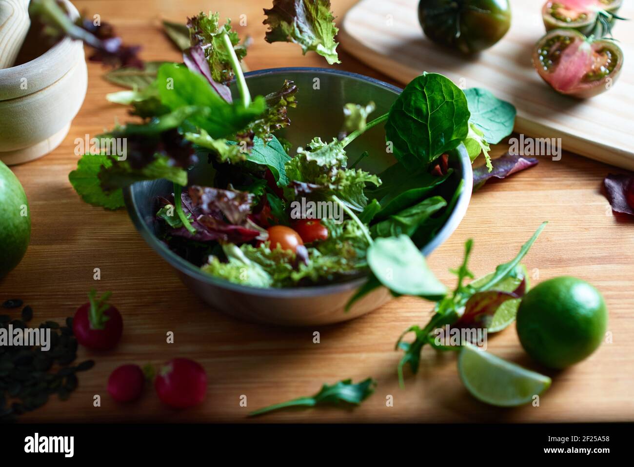 Laitue tombant sur une salade dans un bol en métal sur une table en bois dans une cuisine. Dans la photographie gastronomique, vous pouvez voir des radis, des graines, des planches à découper Banque D'Images
