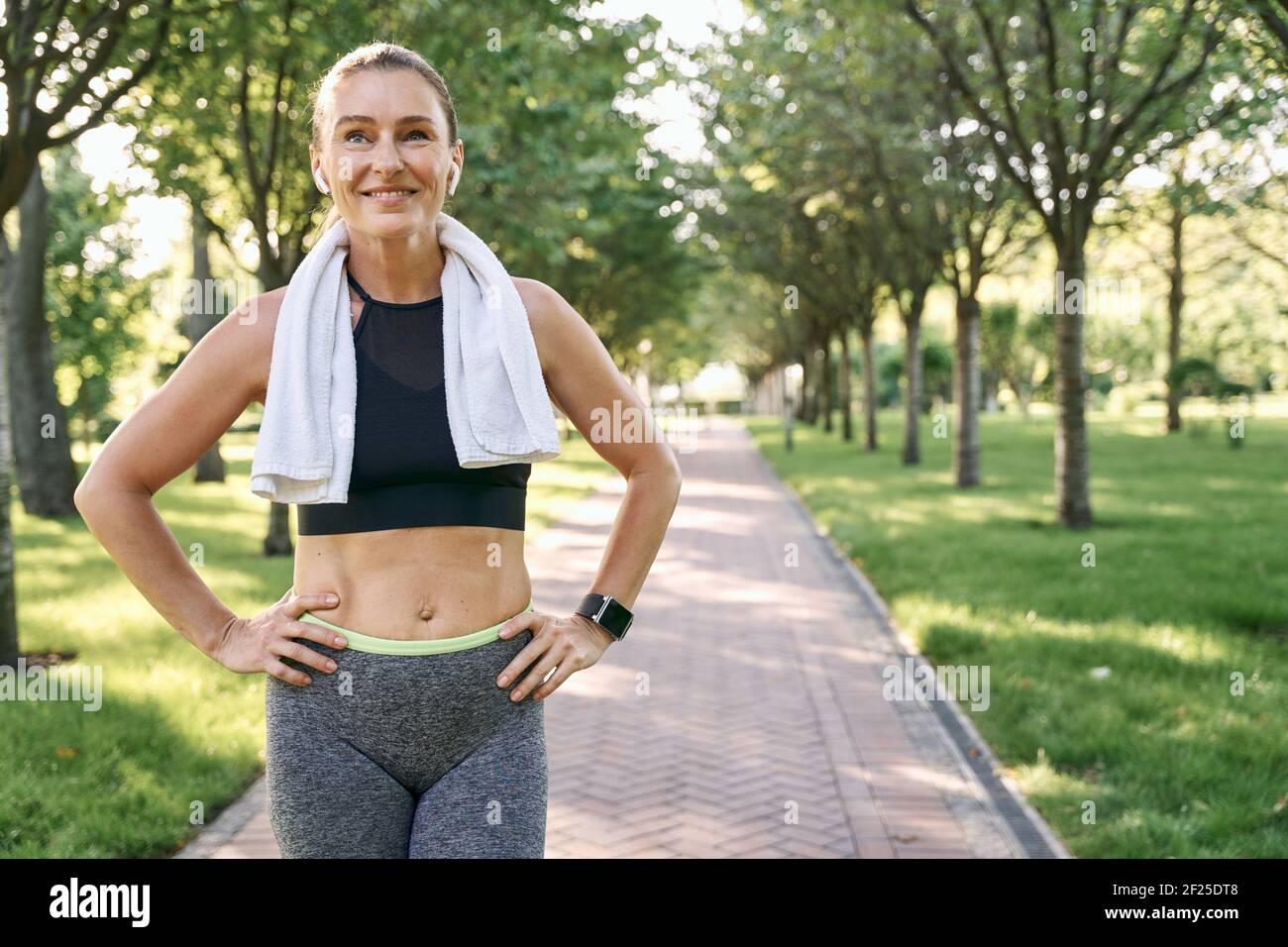 Restez actif. Bonne femme sportive dans des vêtements de sport et des écouteurs sans fil souriant, debout avec une serviette autour de son cou après avoir fait du jogging dans un parc vert Banque D'Images