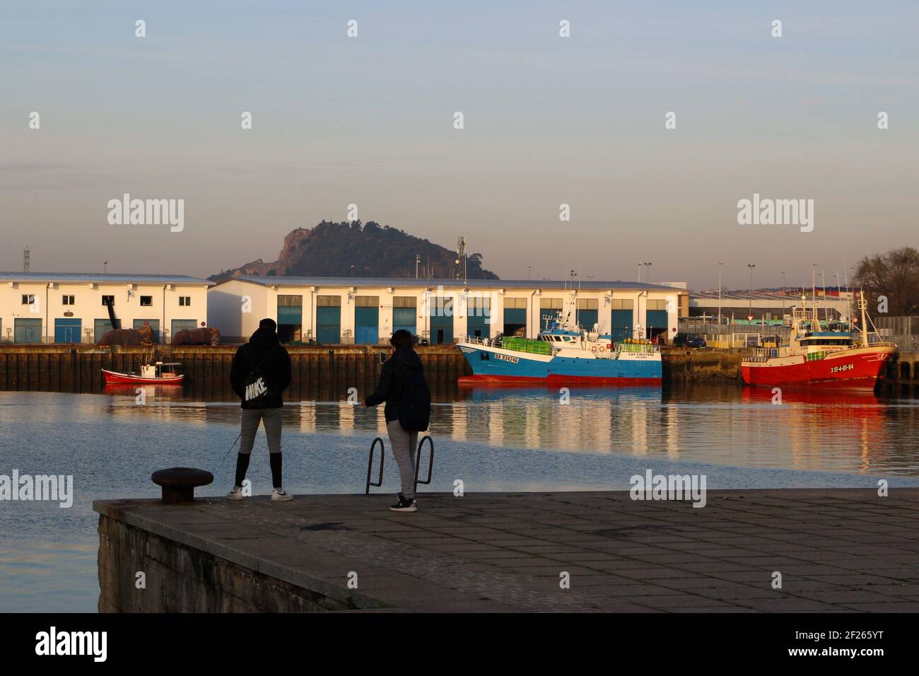 Deux jeunes hommes pêchant dans le Dock de Maliano avec le chalutier français Cap Finistère et le chalutier espagnol Sukari Primero amarrés sous le soleil d'hiver du matin Banque D'Images