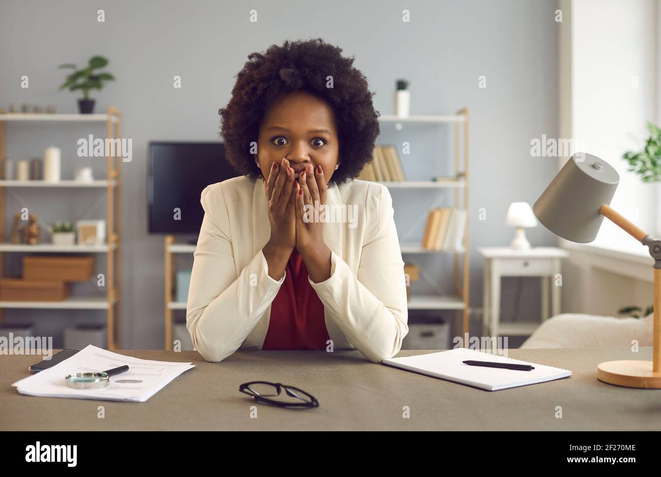 Portrait d'une femme afro-américaine choquée couvrant la bouche avec les mains asseyez-vous au bureau Banque D'Images
