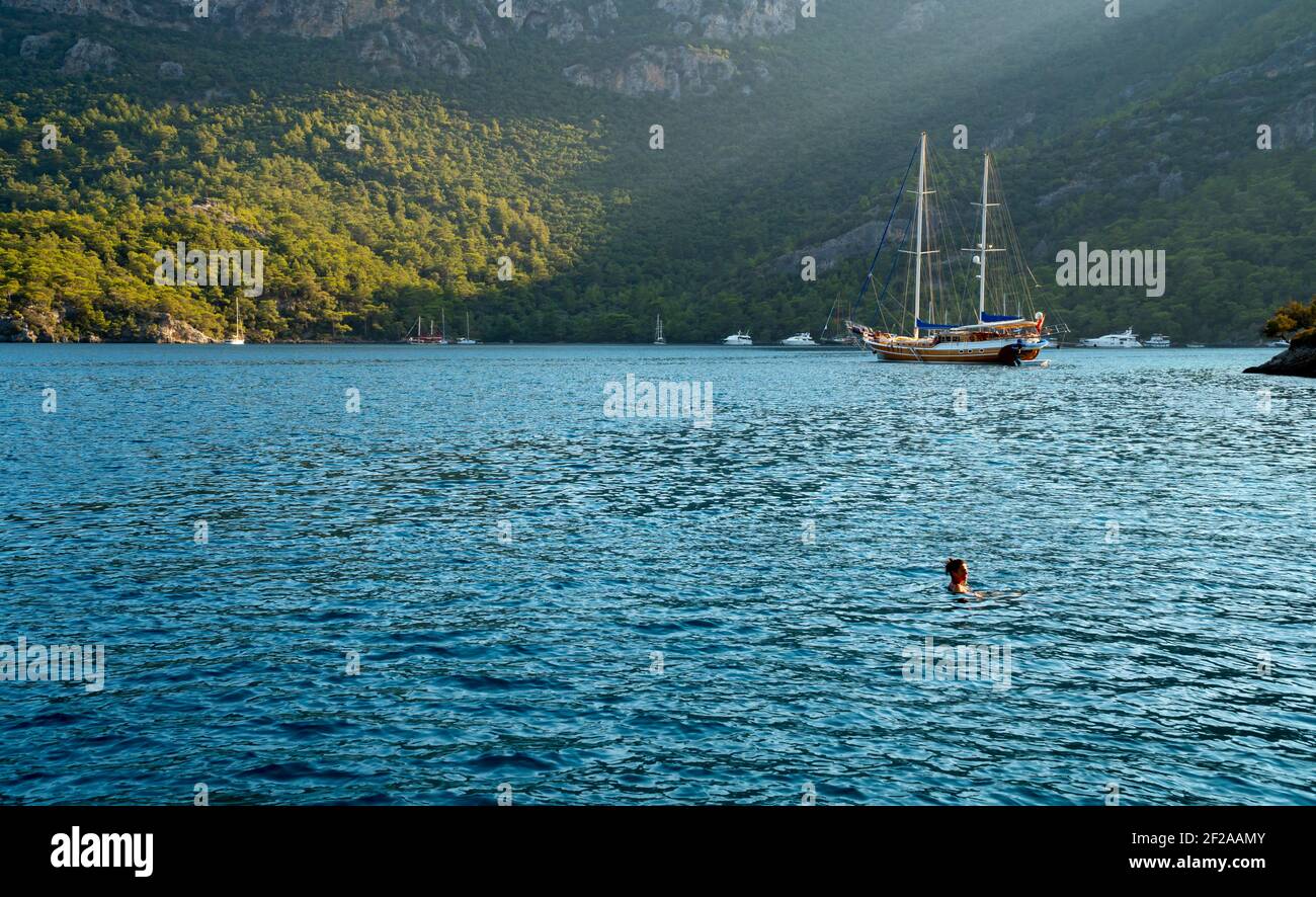 Concept d'été : un yacht turc de gullet ancré à la mer Égée avec un faisceau de soleil en arrière-plan. Une seule femme nageant dans le lac. Contraste vert - bleu Banque D'Images