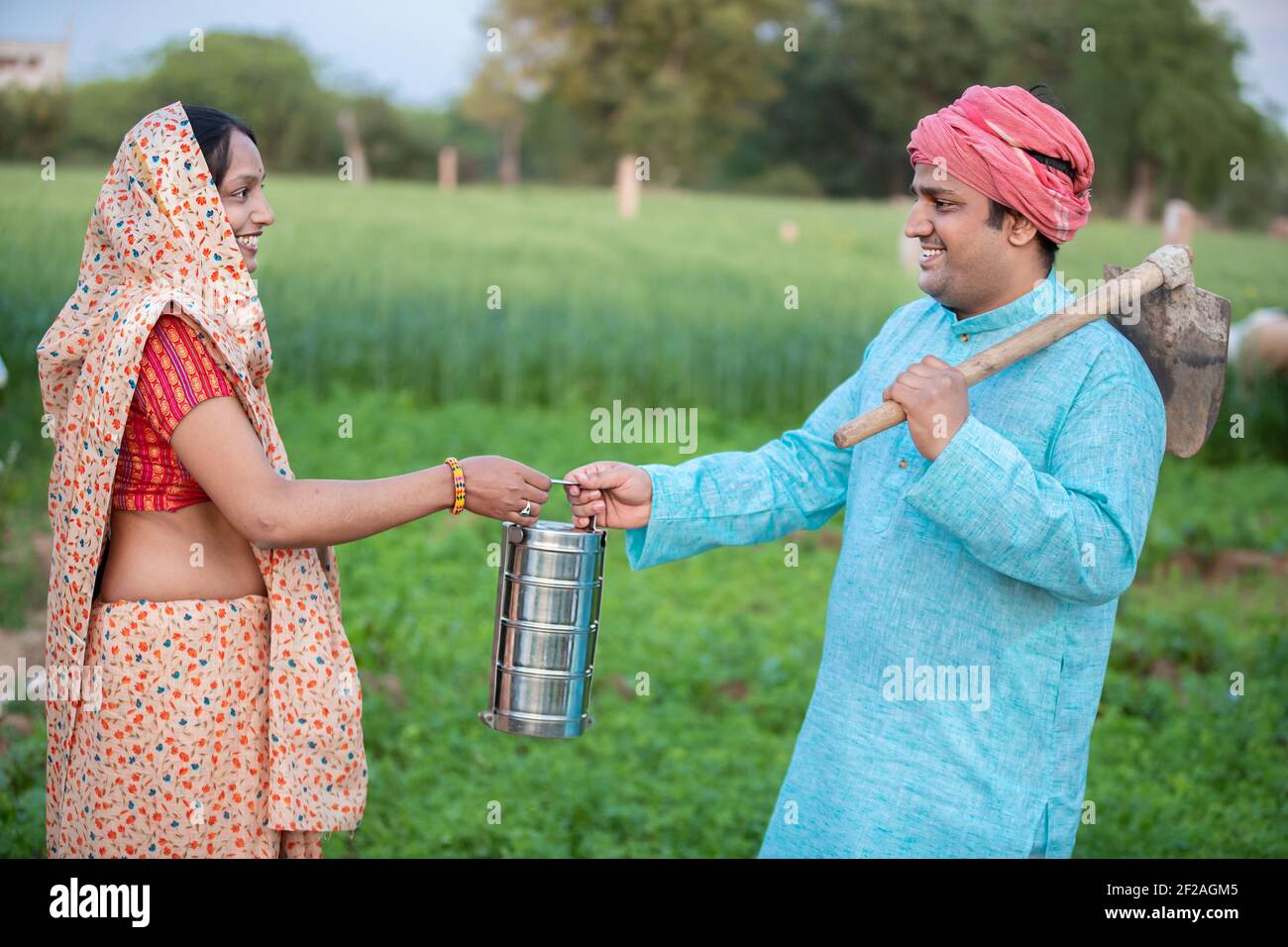 Bonne femme indienne traditionnelle portant sari donnant une boîte de tiffin à son mari dans le domaine agricole, heureux couple fermier tenant une boîte à lunch. Banque D'Images