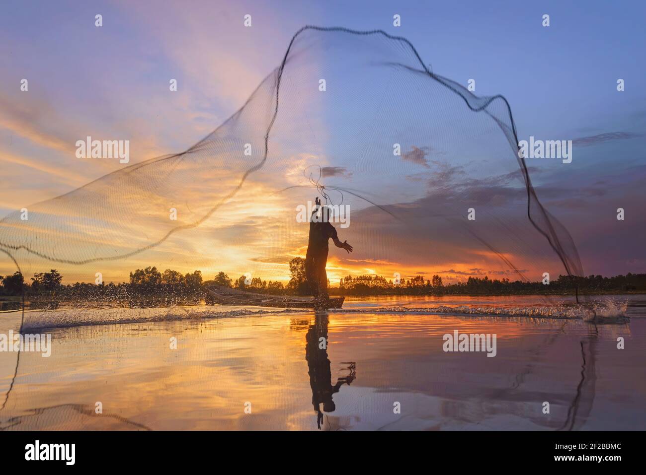 Silhouette d'un pêcheur debout dans un bateau qui jette un filet de pêche au coucher du soleil, Thaïlande Banque D'Images