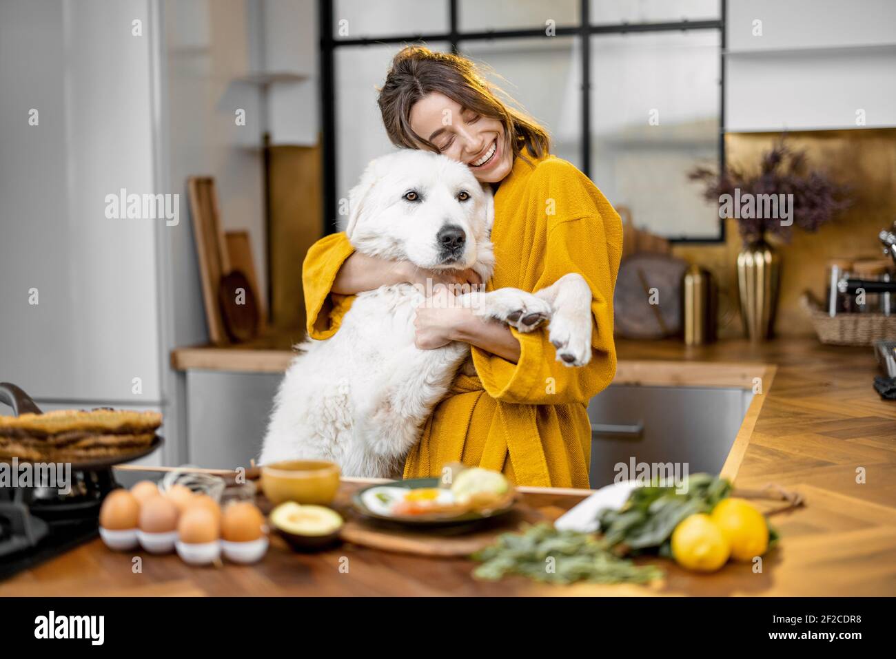 Une jeune femme joue avec son énorme chien blanc tout en ayant un petit-déjeuner pendant une heure du matin sur la cuisine à accueil Banque D'Images