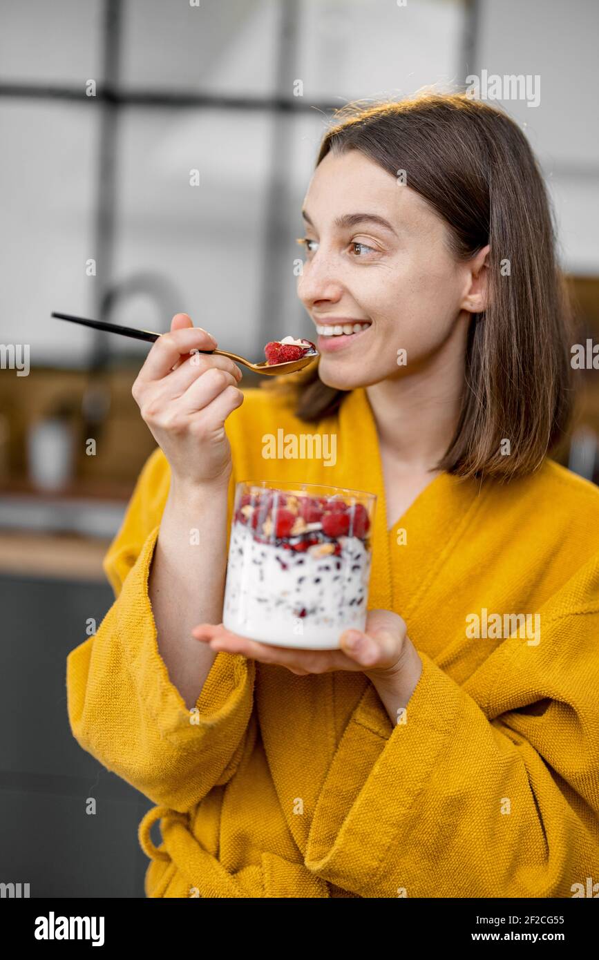 Une jeune femme en peignoir jaune prend un petit déjeuner sain de céréales avec yaourt et baies dans le bol de la cuisine à la maison le matin. Un look souriant et agressif. Banque D'Images