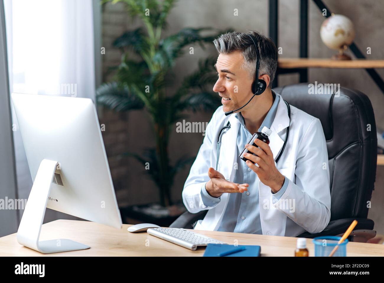 Un médecin sympathique, portant un blouse de laboratoire et un casque,  parlant par appel vidéo avec un ordinateur et un patient, consultation en  ligne. Concept d'aide médicale à distance Photo Stock -